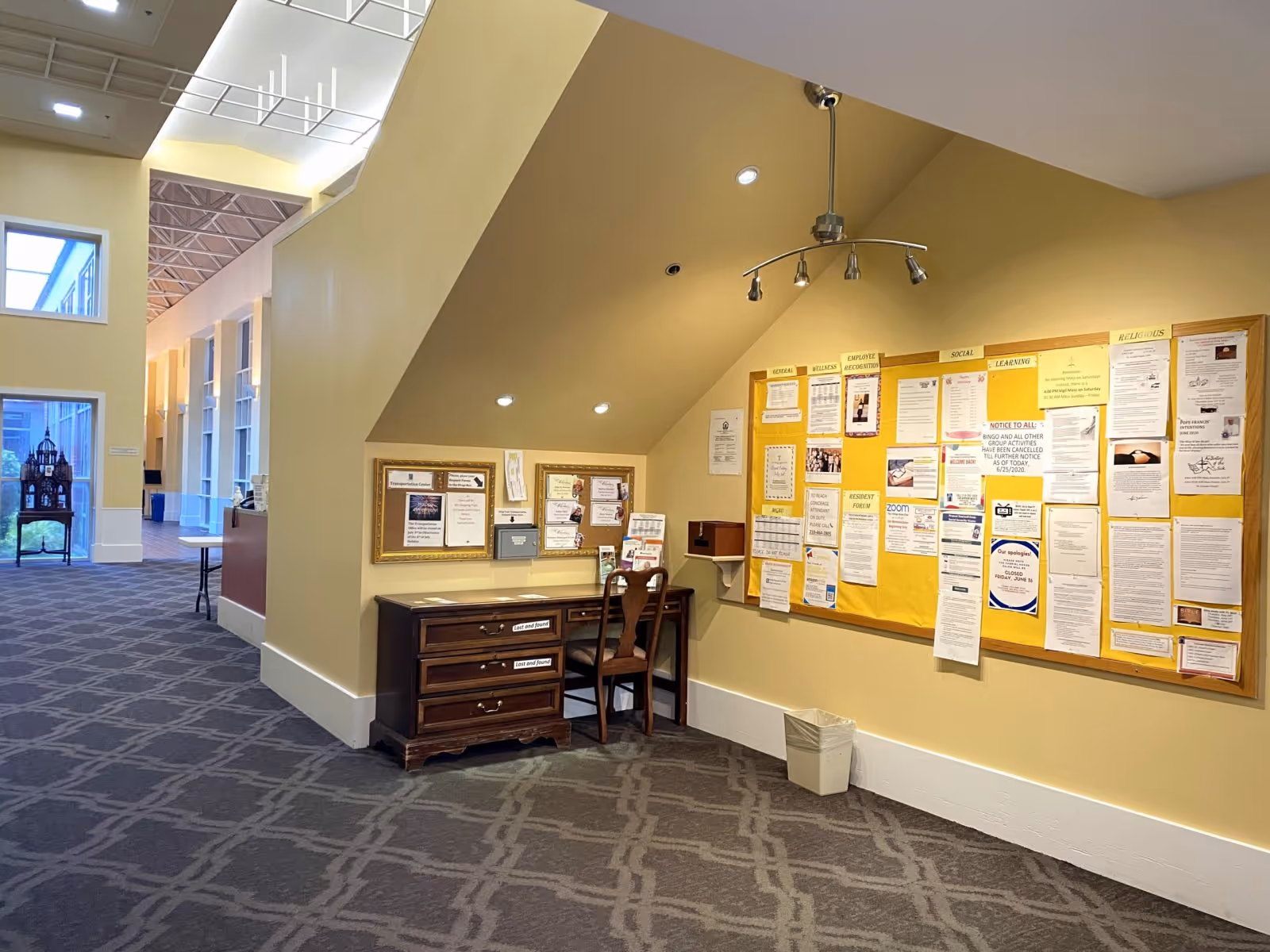 Carpeted interior hallway with a small desk and chair beneath a sloped ceiling and a bulletin board filled with notices.