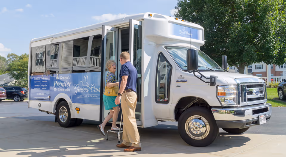 An elderly woman is stepping onto a white shuttle bus with blue signage that reads 'Lake Geneva's Premier 55+ Community Geneva Crossing.' A man is standing nearby assisting her. The scene is outdoors on a sunny day with trees and parked cars in the background.
