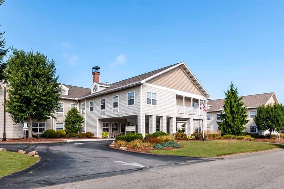 Two-story beige senior living building with a covered entrance, landscaped lawns and a curved driveway under a clear blue sky.