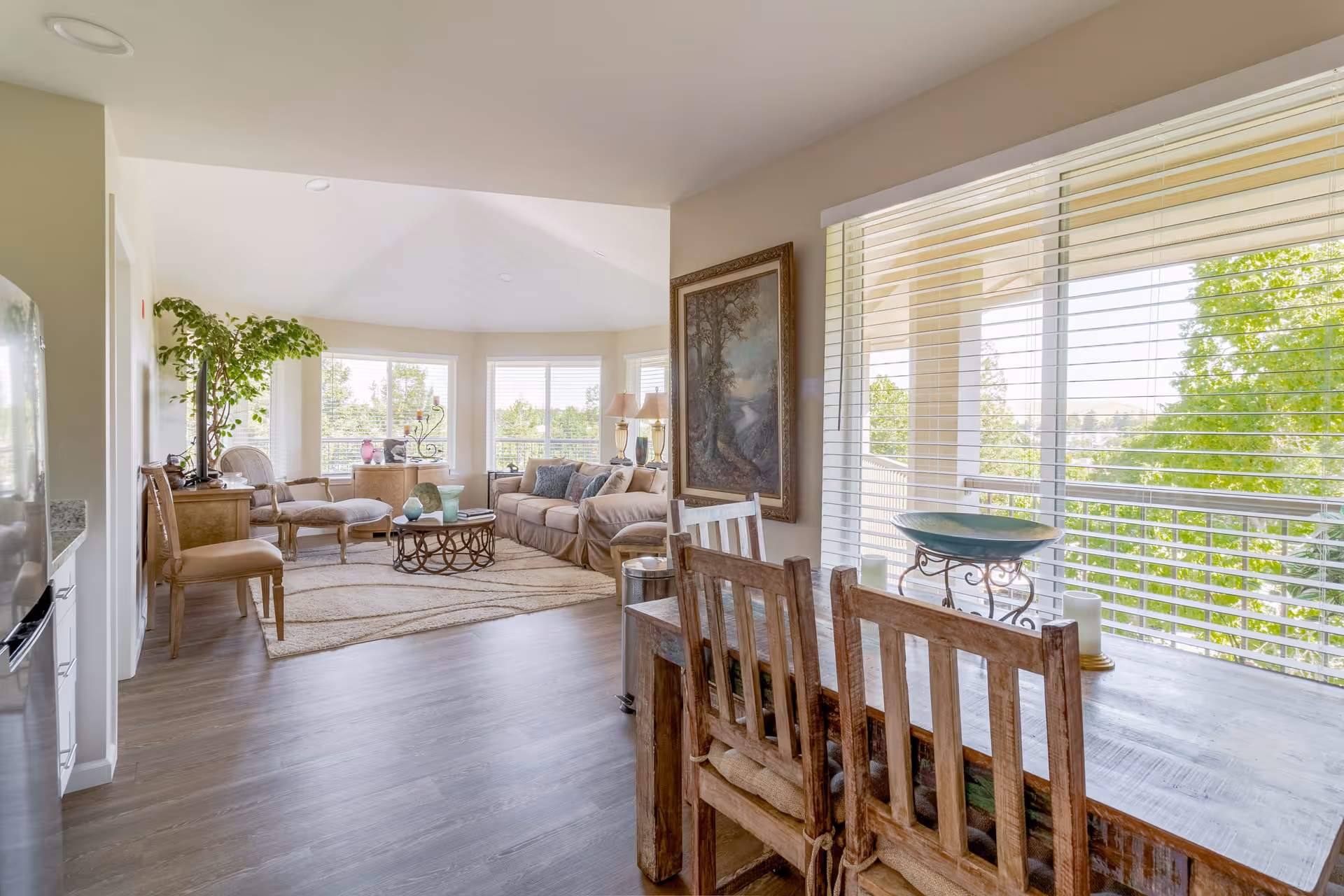 A bright and spacious living area in a retirement community featuring a wooden dining table with chairs near large windows with blinds. The living room has a beige sofa, armchairs, a coffee table, a large rug, and decorative items including a potted plant and framed artwork on the wall. Outside the windows, green trees and a balcony are visible.