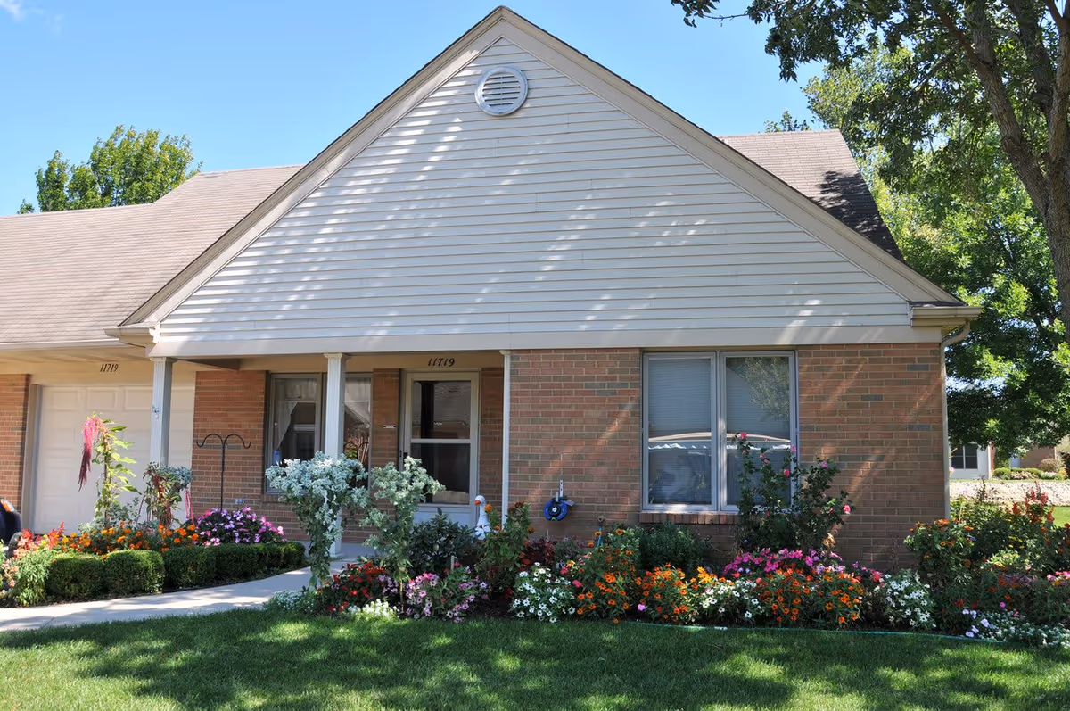 Exterior view of a single-story brick and siding building with a gable roof, a front porch with two white columns, a window with blinds, and a garden bed filled with colorful flowers and greenery in front of the building under a clear blue sky.