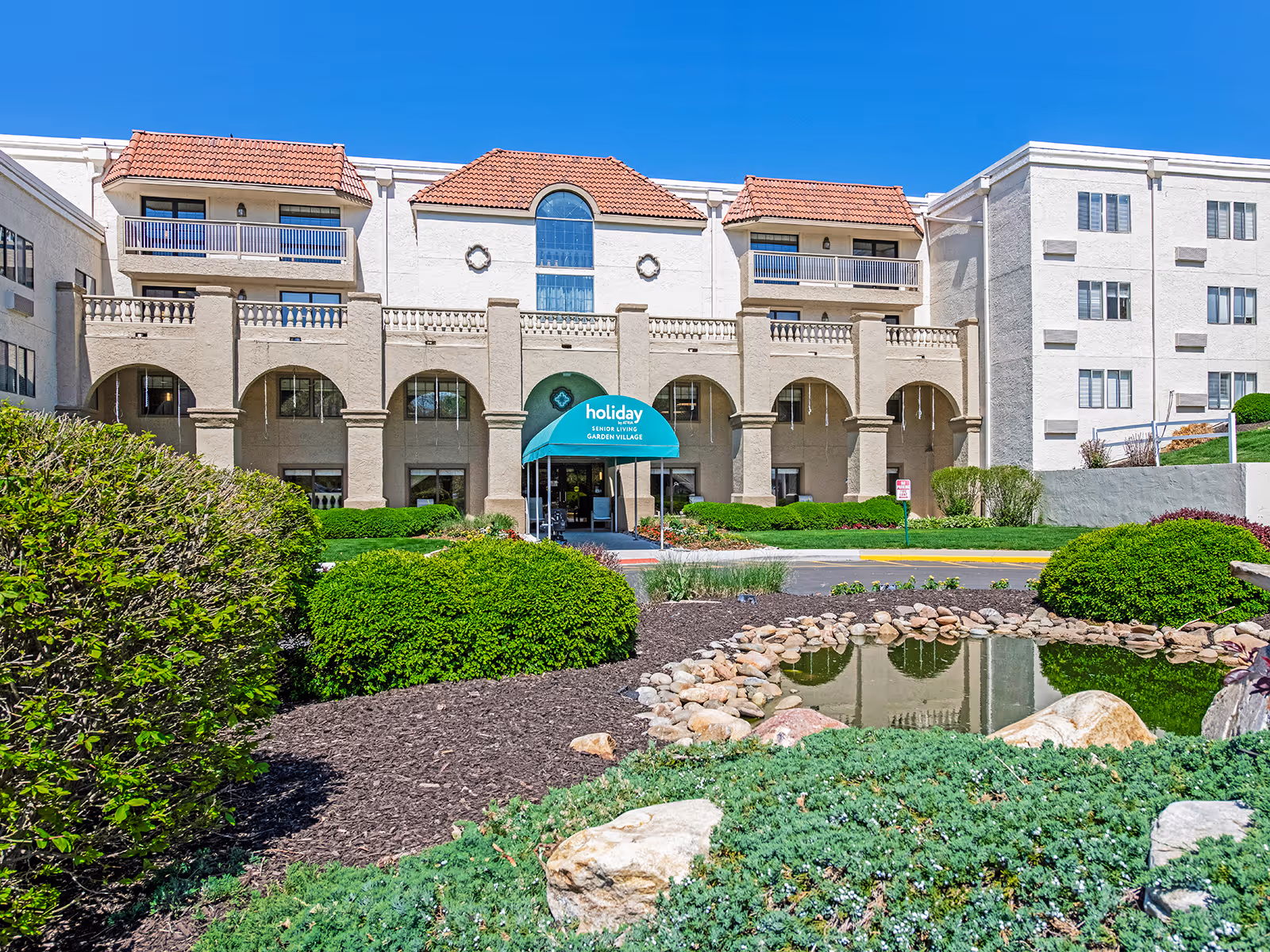 Exterior view of Garden Village senior living facility with landscaped pond and arched entrance.