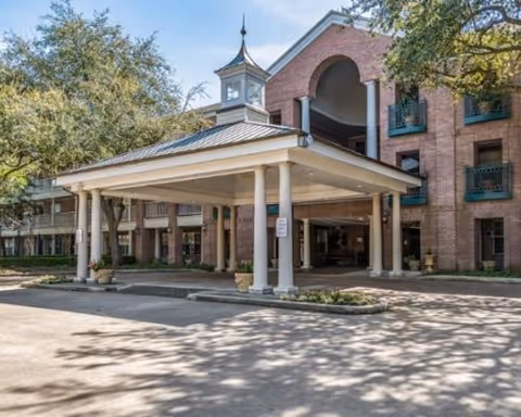 Entrance of a multi-story brick building with a covered drop-off area supported by white columns, surrounded by trees and greenery under a clear blue sky.