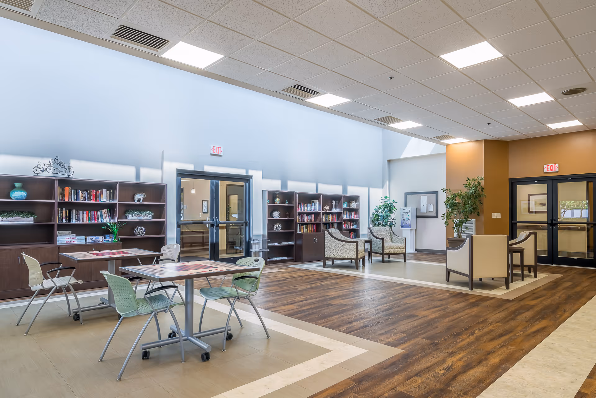 A spacious senior living common area with wooden flooring and beige carpeted sections. The room features several tables with chairs, bookshelves filled with books and decorative items, and comfortable armchairs arranged near potted plants. Large windows and ceiling lights provide ample lighting, and there are double glass doors with exit signs above them.