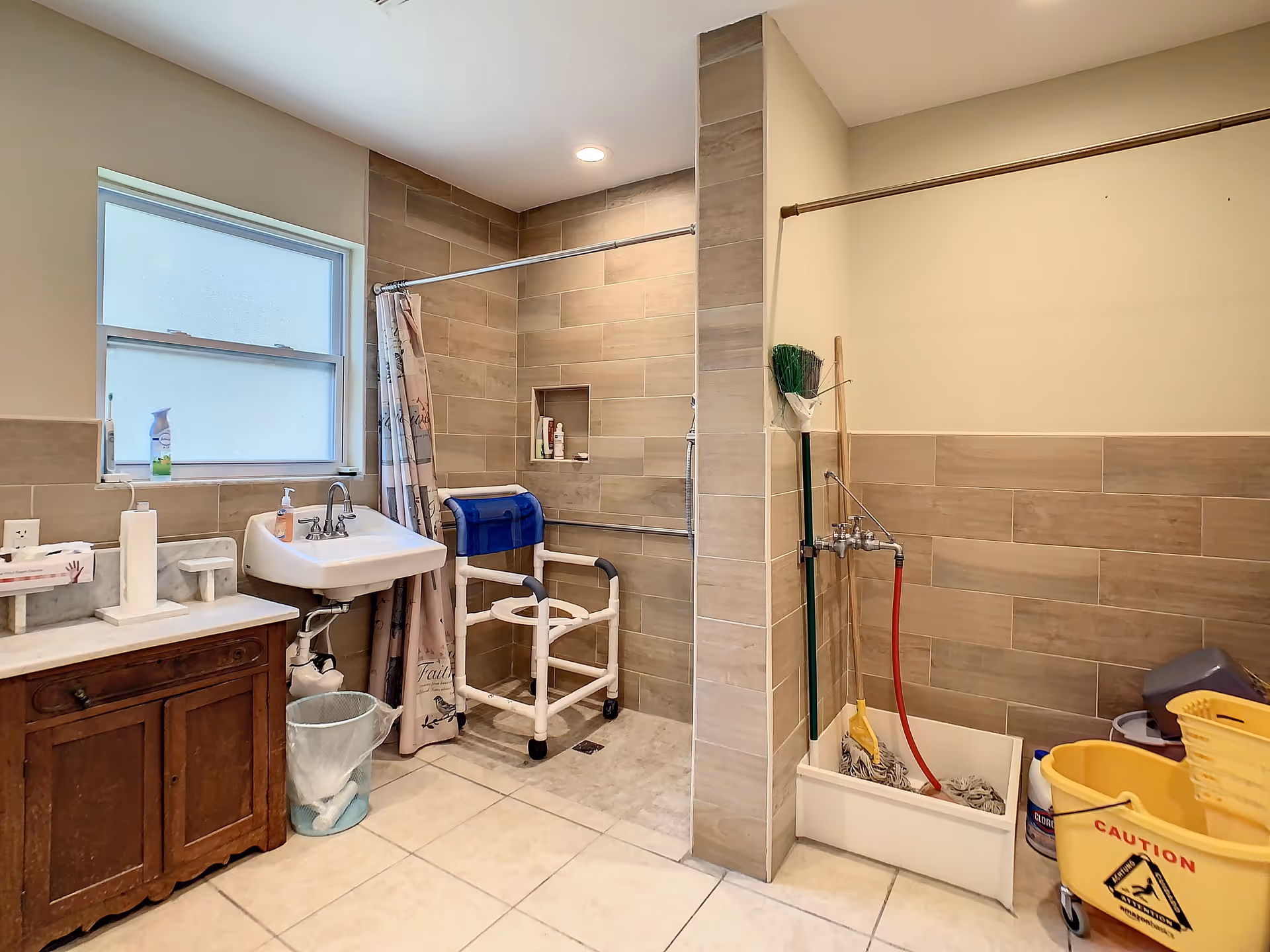 A bathroom with beige tiled walls and floor, featuring a walk-in shower with a white shower chair and a shower curtain. There is a white sink mounted on the wall with soap and cleaning supplies on the counter next to it. On the right side, there is a cleaning station with a mop, broom, bucket, and caution sign.