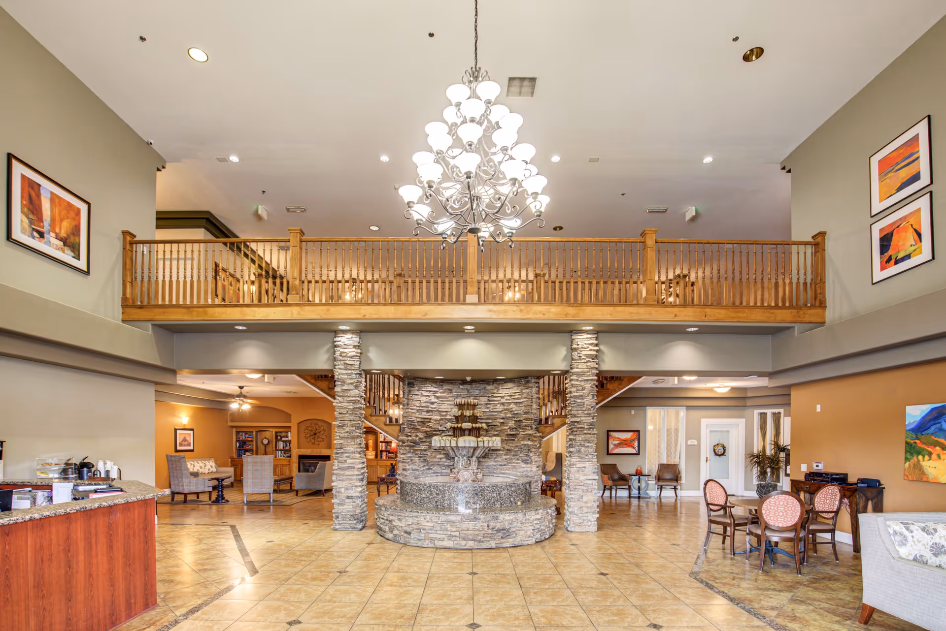 Spacious senior living facility lobby with a large chandelier hanging from a high ceiling, a stone fountain centerpiece, wooden balcony railing on the second floor, and seating areas with chairs and tables on either side. The walls are decorated with colorful framed artwork.