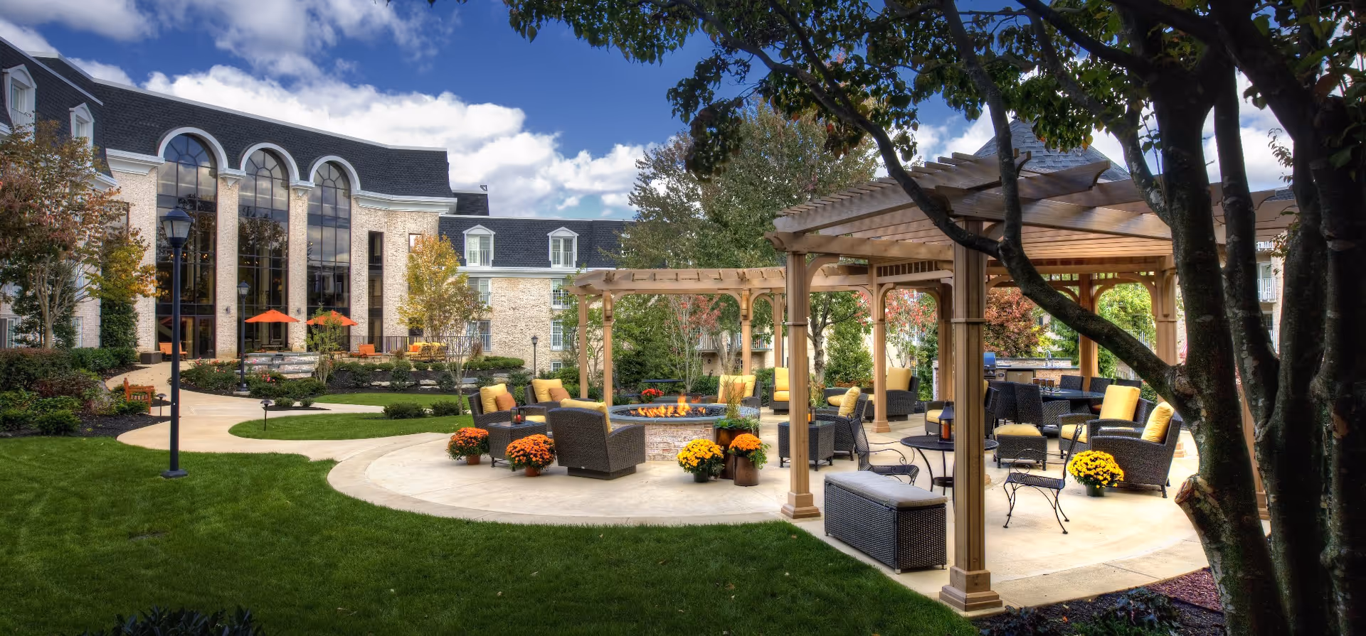 Outdoor courtyard with pergolas, seating areas, potted flowers and a central fire pit in front of a large building.