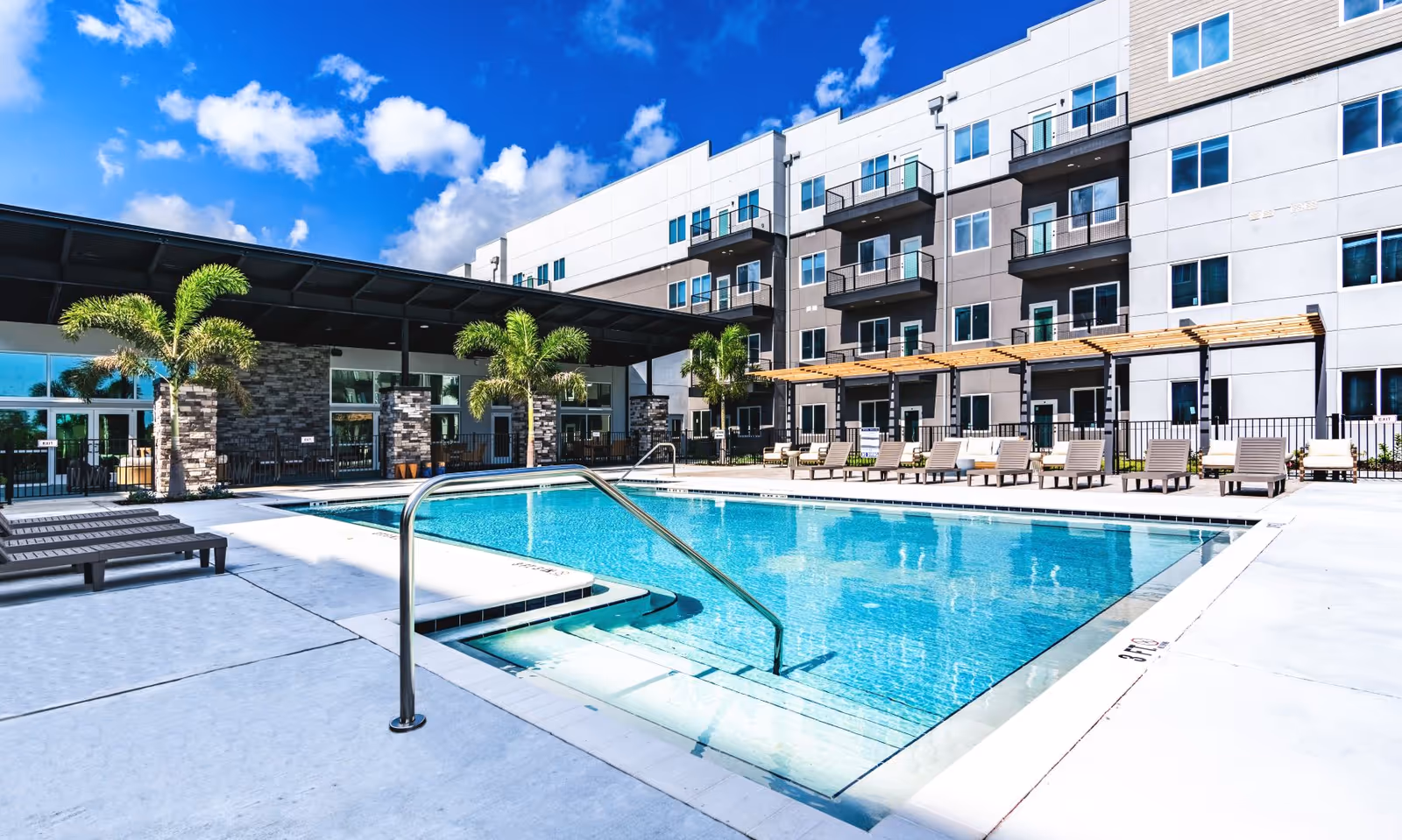 Outdoor swimming pool with lounge chairs and palm trees in front of a multi-story residential building under a blue sky.