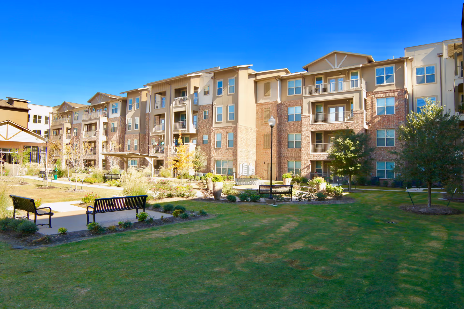 Exterior view of a multi-story senior living facility building with balconies, surrounded by a well-maintained lawn, trees, shrubs, and benches under a clear blue sky.