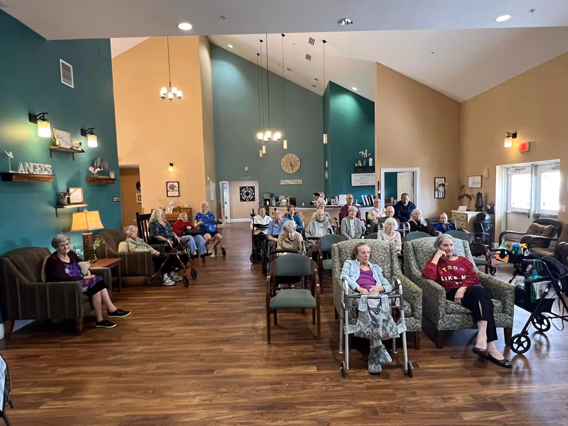 A large common area in a senior living facility with many elderly residents seated in chairs and wheelchairs facing forward. The room has high ceilings, teal and beige walls, wooden floors, and several light fixtures. There are shelves with decorations on the left wall and a clock on the far wall. Some residents are sitting in armchairs near the front, while others are seated in rows of chairs behind them.