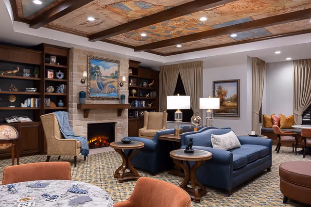 A cozy living room area in a senior living facility featuring a blue sofa with a white pillow, two beige armchairs, wooden side tables with lamps, a stone fireplace with a painting above it, built-in dark wood bookshelves filled with books and decorative items, and a round table with orange chairs in the foreground. The ceiling has a decorative painted design, and the room is warmly lit with recessed lighting.