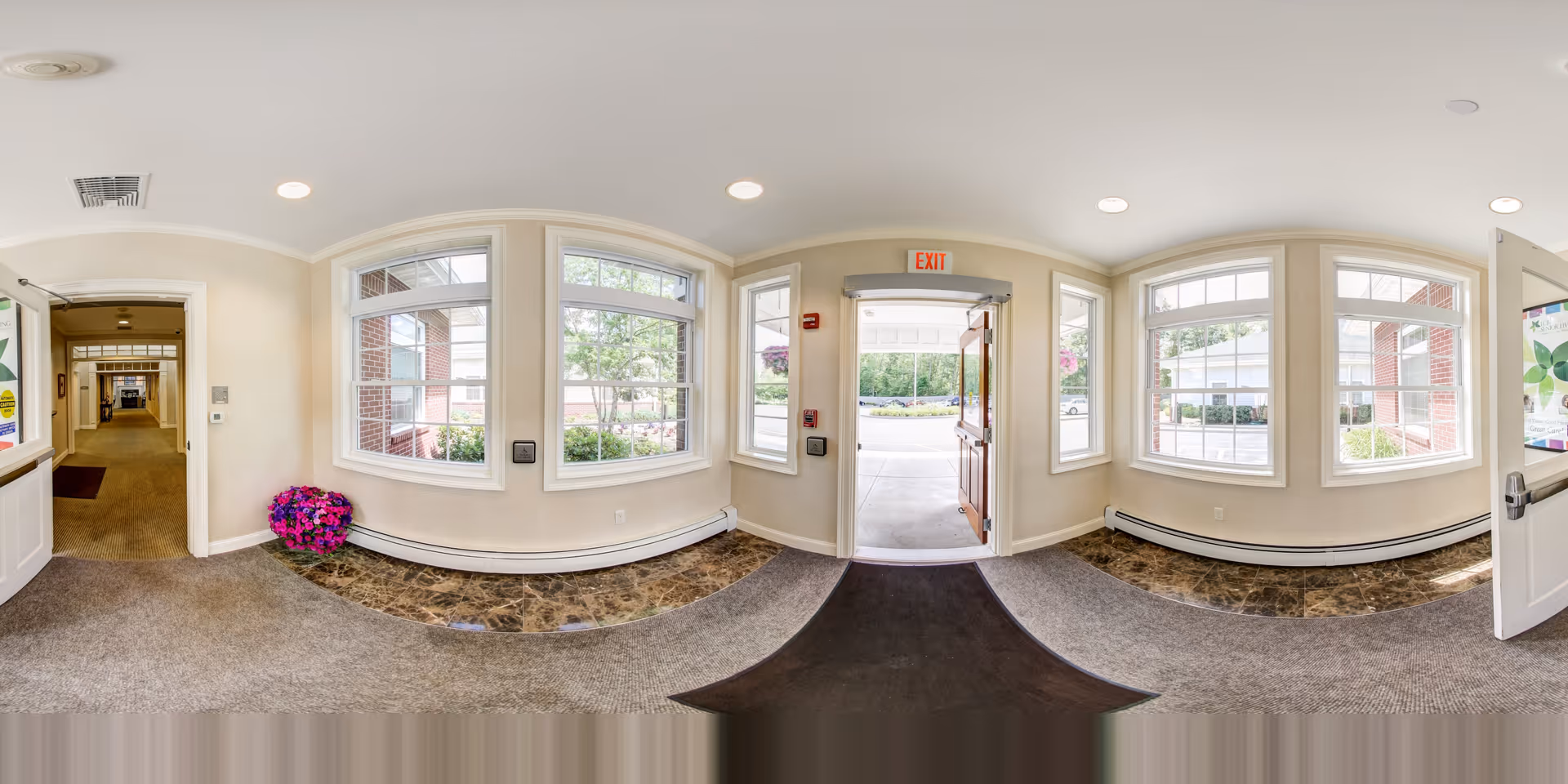 Interior view of an entrance area in The Lighthouse at Lincoln facility showing a door open to the outside, large windows letting in natural light, beige walls, carpeted and tiled flooring, and a hallway leading further inside the building.