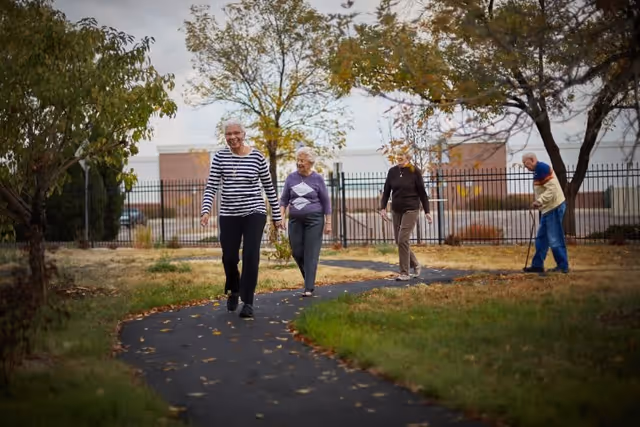 Four elderly people walking along a paved path in an outdoor garden area with trees and grass, with a fence and buildings in the background.