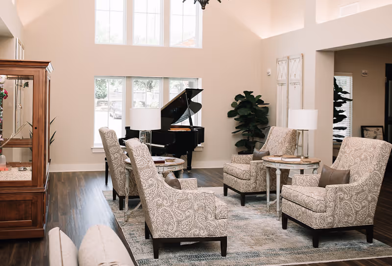 A bright and spacious living room area with four patterned armchairs arranged around two small round tables with lamps. A black grand piano is positioned near large windows letting in natural light. There is a wooden cabinet on the left and a tall green plant near the wall. The room has light-colored walls and wood flooring with a large area rug.