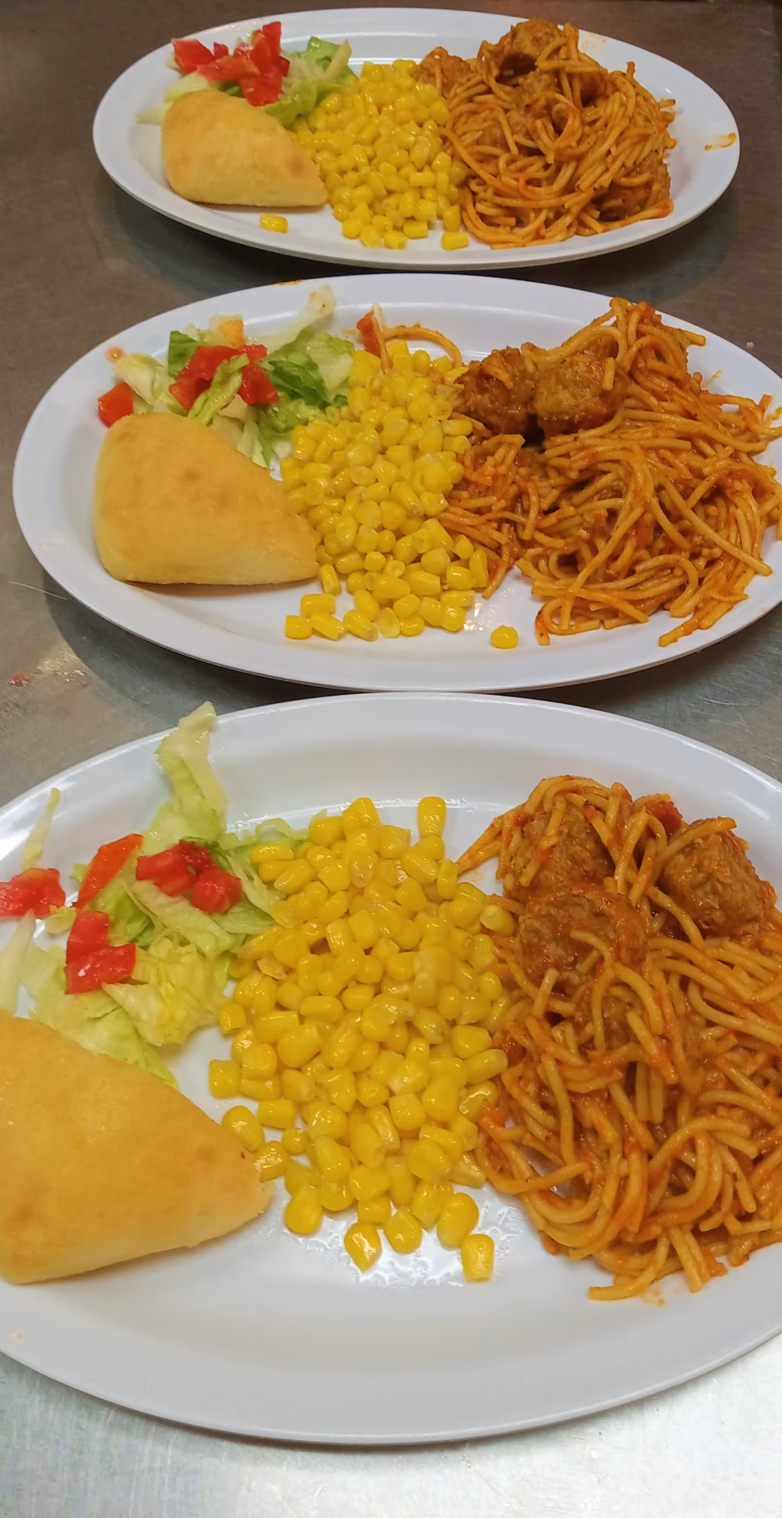 Three white oval plates each containing a serving of spaghetti with meatballs in tomato sauce, a portion of yellow corn, a small salad with lettuce and diced red bell peppers, and a piece of bread, arranged on a metallic surface.