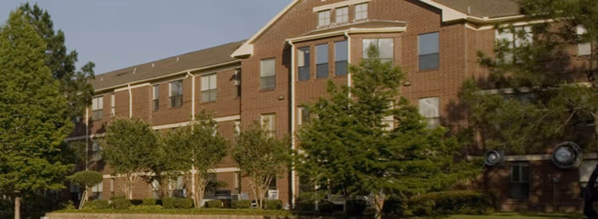 Exterior view of a three-story brick building with multiple windows, surrounded by green trees and shrubs under a clear sky.