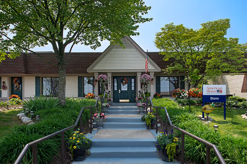 Front exterior view of American House Stone facility with a central entrance featuring steps and handrails, surrounded by green plants, flowers, and trees under a clear blue sky.