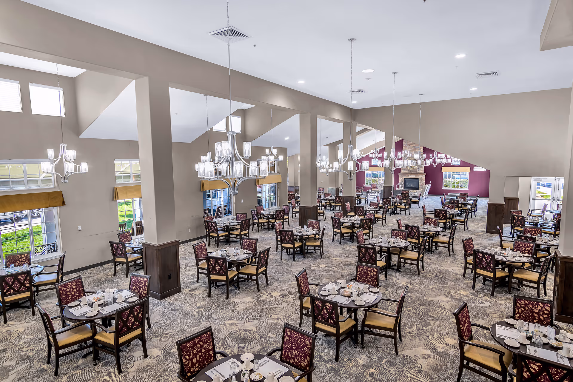 Spacious senior living dining room with many set tables, chandeliers, and high ceilings.