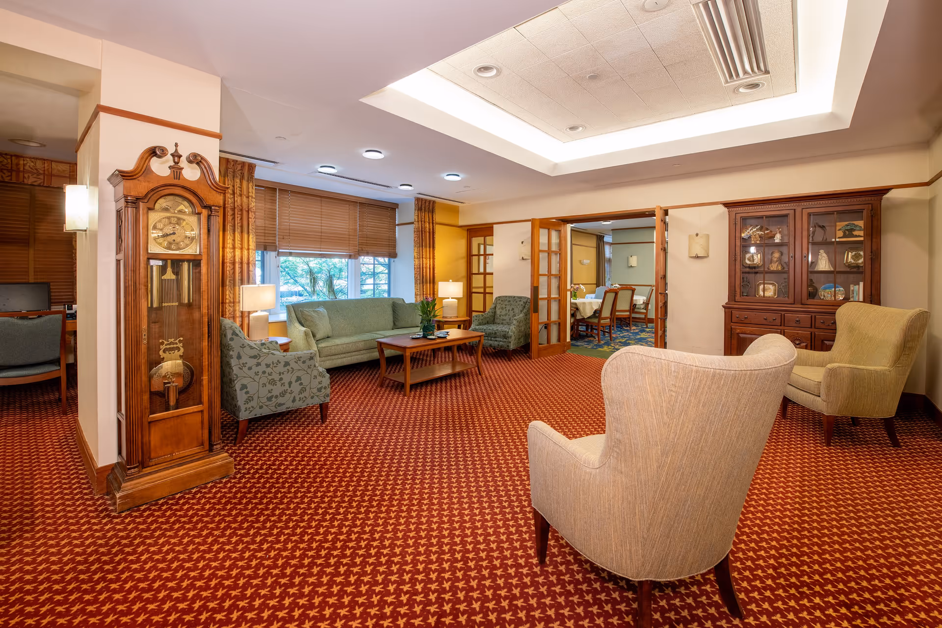 A cozy living room area in a senior living facility with a red patterned carpet, beige armchairs, a green sofa, and a wooden coffee table. There is a tall wooden grandfather clock on the left side and a wooden cabinet with glass doors on the right. The room has large windows with wooden blinds and yellow curtains, and French doors leading to a dining area with tables and chairs.