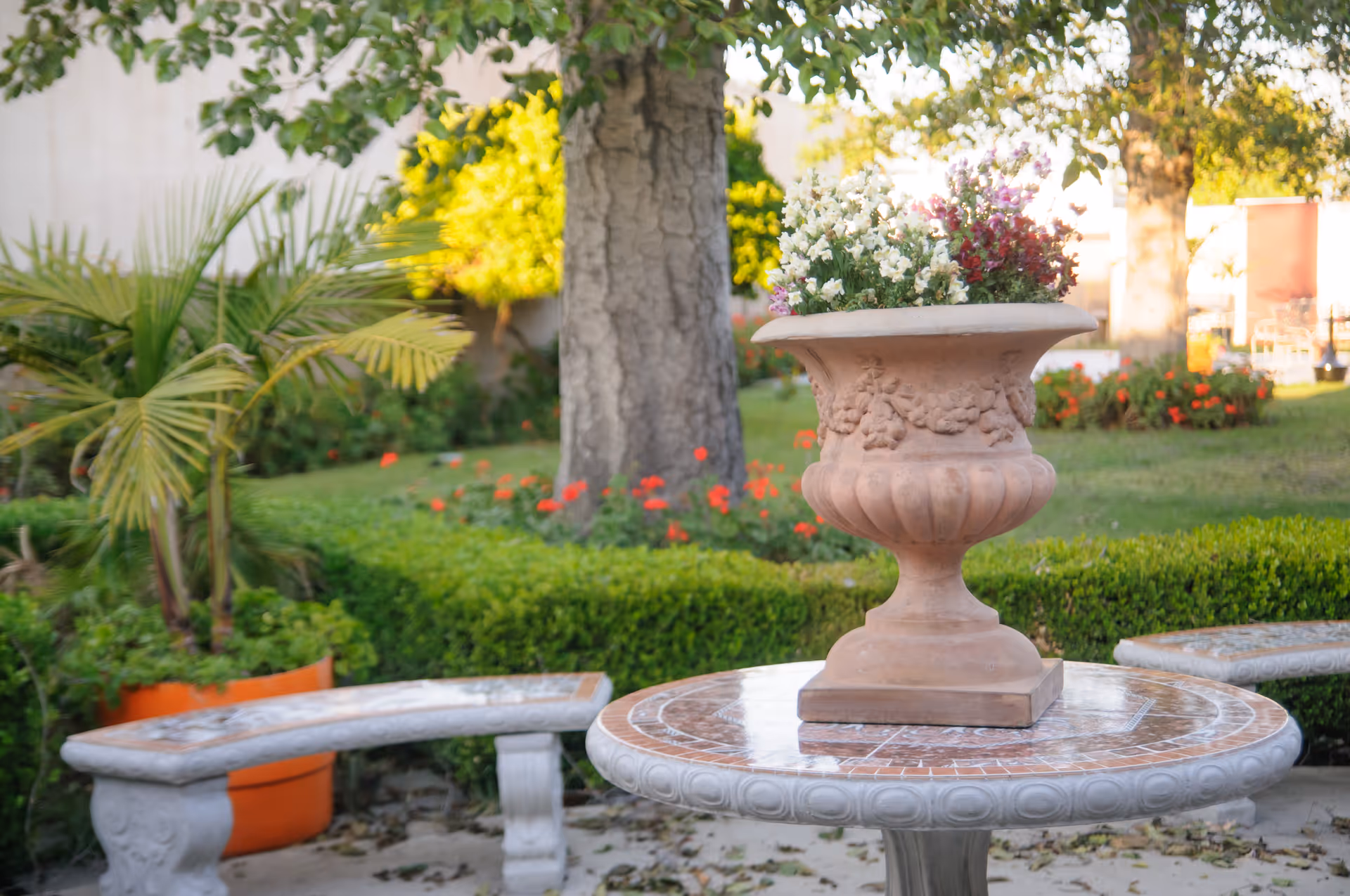 A decorative outdoor garden scene featuring a round stone table with a large ornate planter filled with white and purple flowers. Surrounding the table are curved stone benches. In the background, there are green bushes, a large tree trunk, and other plants in a well-maintained garden area.
