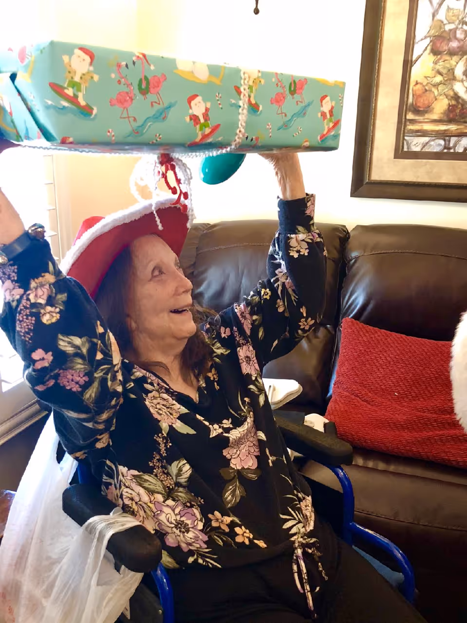 An elderly woman wearing a red and white Santa hat and a floral blouse is sitting in a wheelchair indoors. She is smiling and holding a large wrapped gift box above her head. Behind her is a brown leather couch with a red cushion and a framed painting on the wall.