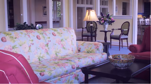 A cozy living room area featuring a floral patterned sofa, a dark wooden coffee table with a decorative bowl, a red armchair, and a side table with a lamp and a vase of flowers. In the background, there are additional chairs and a glimpse into another room.