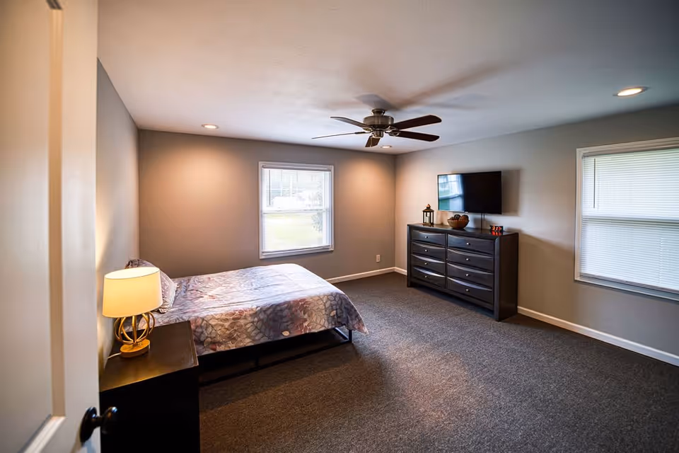 A bedroom with a bed covered in a patterned bedspread, a bedside table with a lamp, a ceiling fan, two windows with blinds, and a dresser with a flat-screen TV mounted above it.