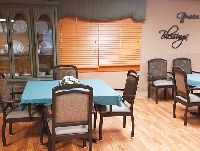 A dining room with wooden floor, two tables covered with teal tablecloths, and several cushioned chairs around them. A china cabinet with plates is visible on the left side, and a window with closed blinds is in the center background. On the right wall, decorative words 'Grace' and 'blessings' are mounted.