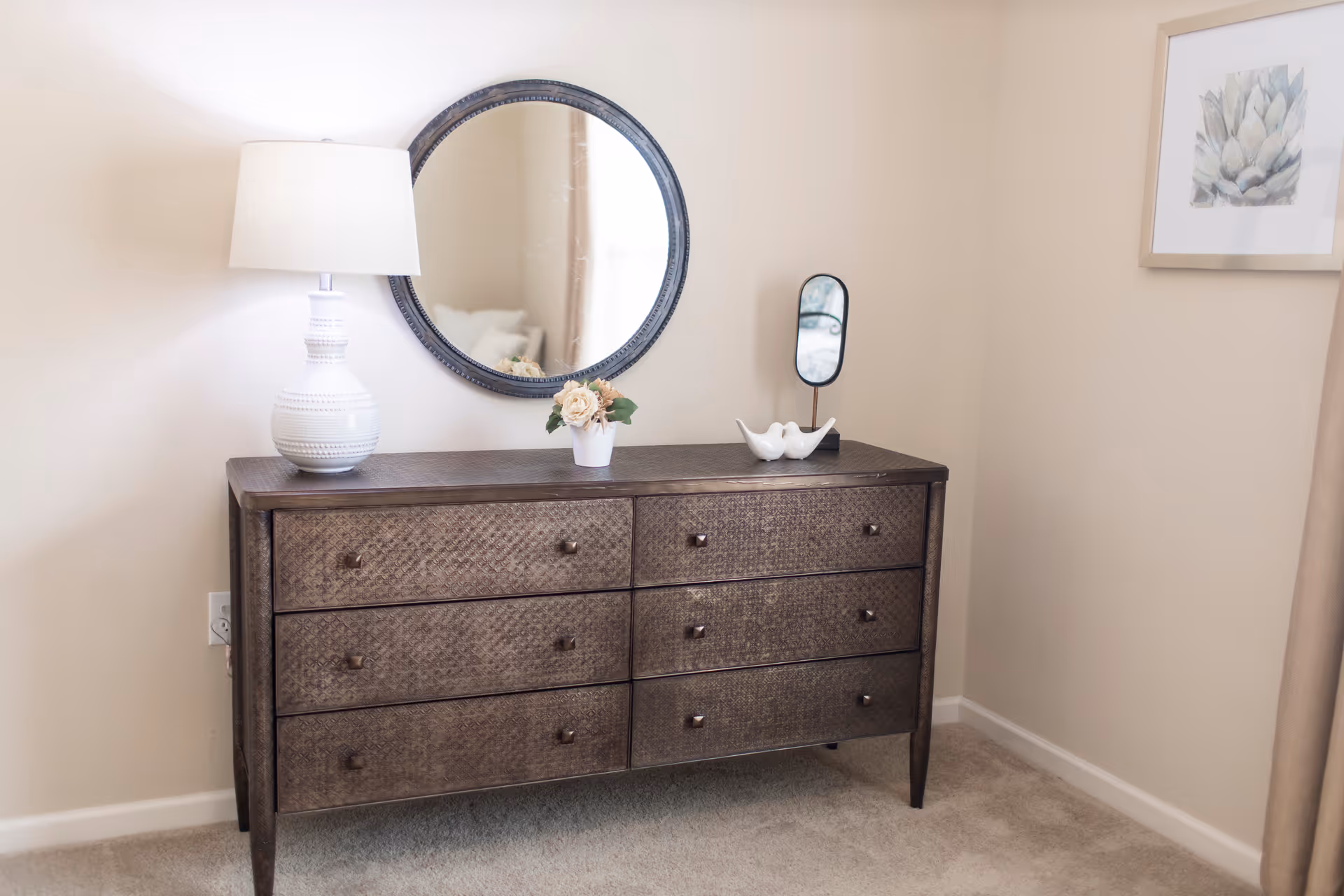 A wooden dresser with six drawers stands against a beige wall. On top of the dresser is a white table lamp, a small potted flower arrangement, and a decorative white bird figurine. Above the dresser hangs a round mirror with a dark frame. To the right, part of a framed floral artwork is visible on the wall.
