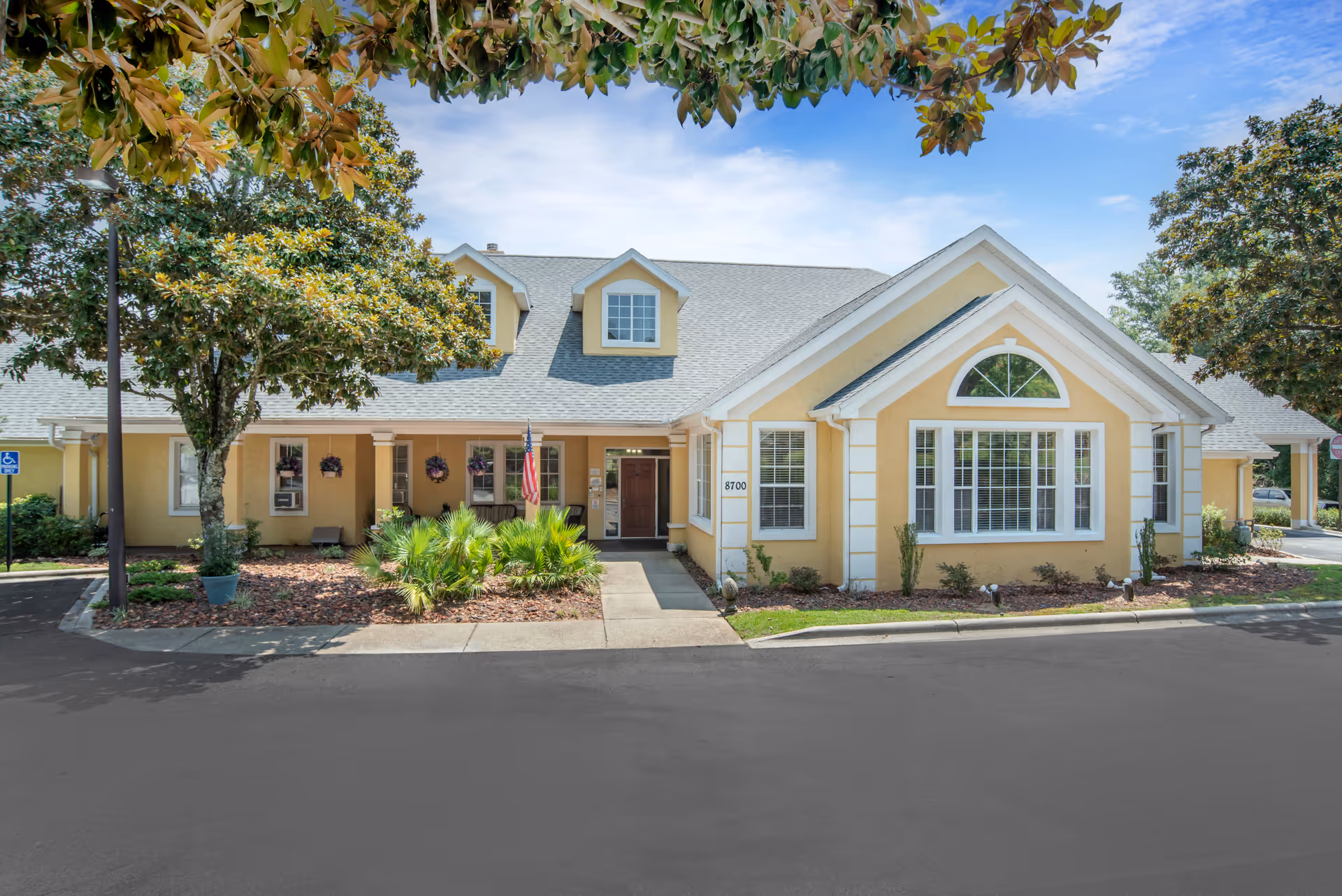 Front exterior of a yellow, gabled senior living building with a central entrance, large windows, landscaping, and an American flag.