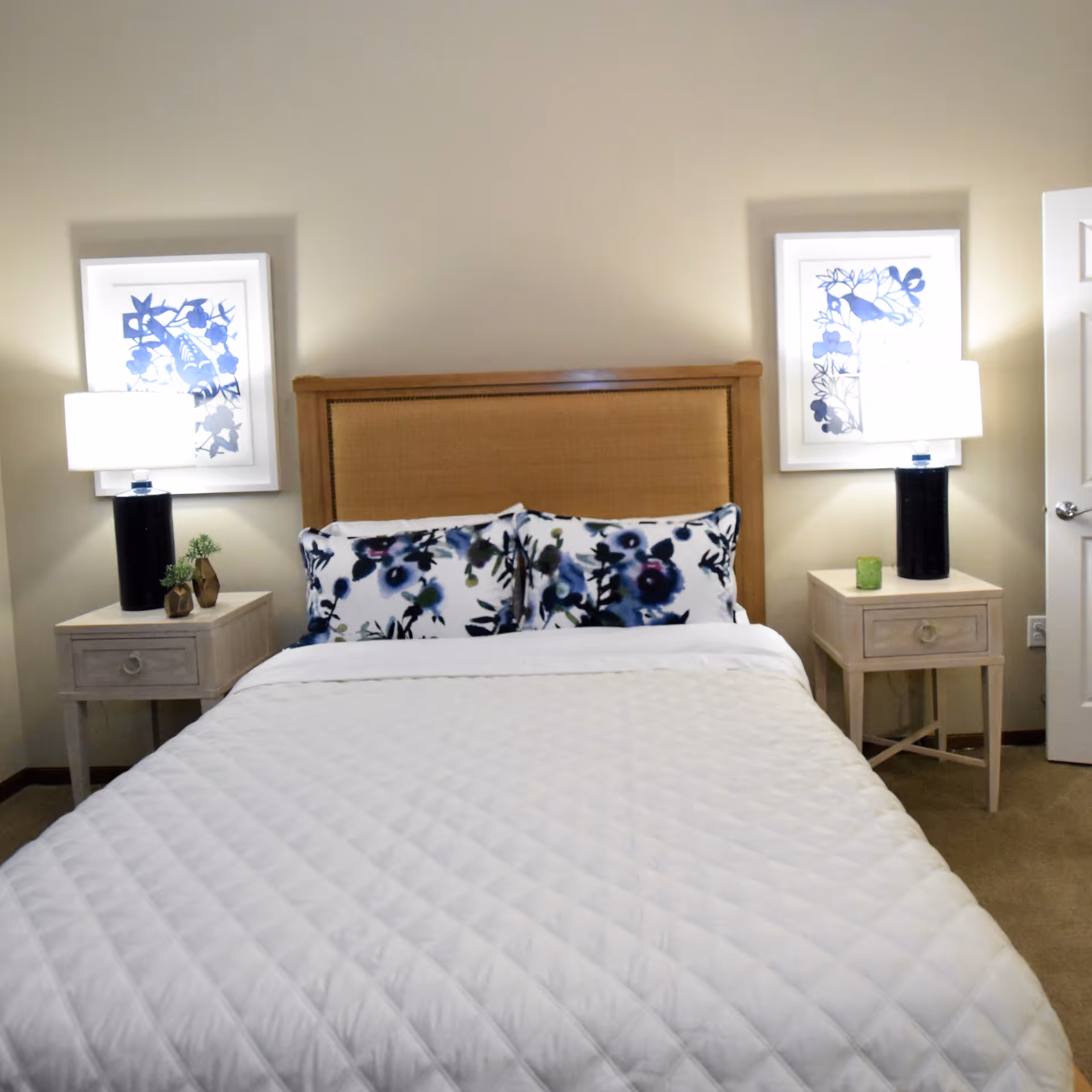 A neatly made bed with a wooden headboard, white quilted bedspread, and two floral-patterned pillows. On each side of the bed are light-colored wooden nightstands with black lamps and framed blue and white artwork hanging above them. The room has beige walls and carpeted floor.