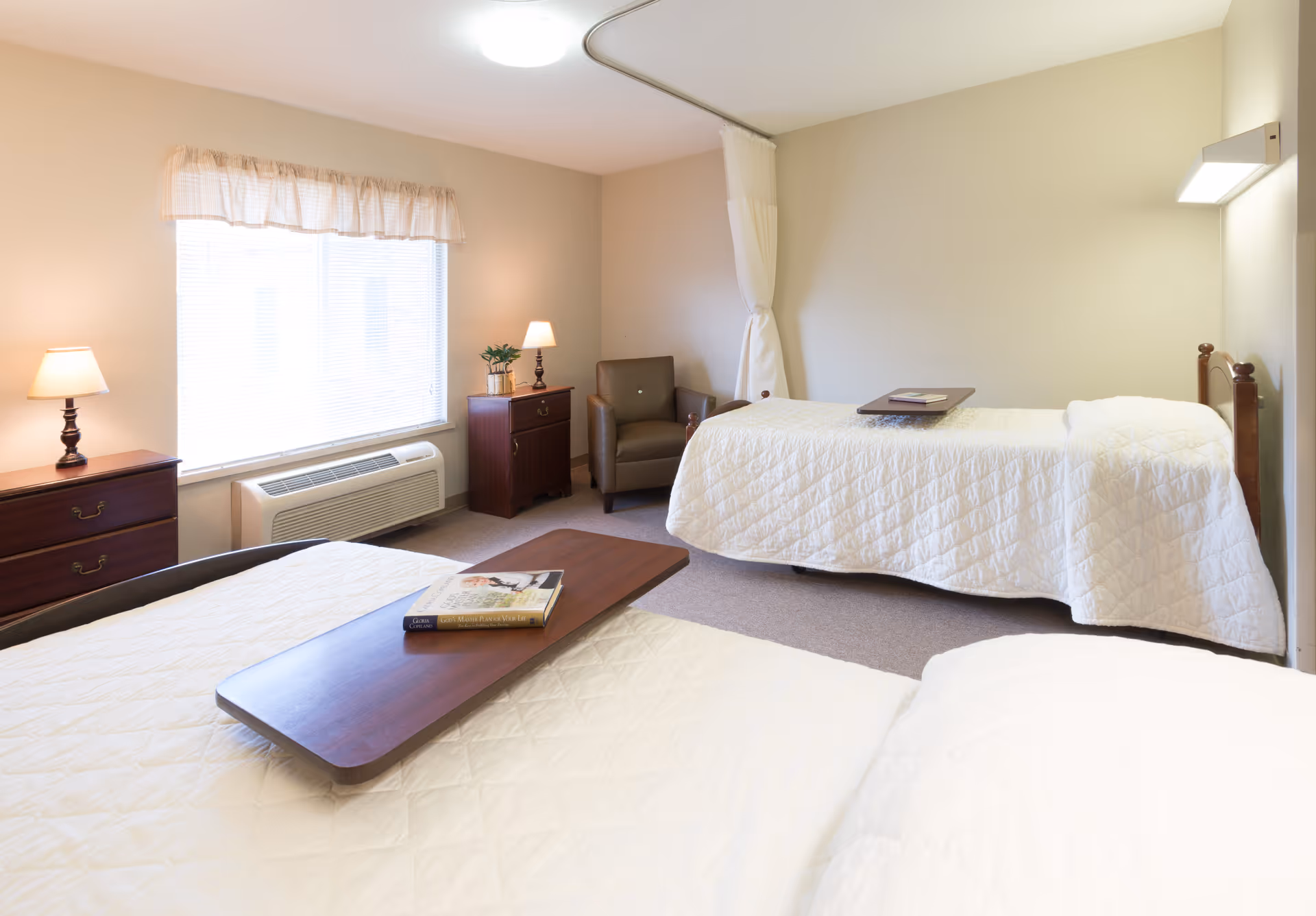 A bright and clean bedroom in a senior living facility with two single beds covered in white quilts, each with a wooden overbed table. The room has beige walls, a window with blinds and a valance, two wooden dressers with lamps, a brown armchair, and a white privacy curtain hanging from the ceiling.
