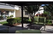 Outdoor seating area with green cushioned chairs and a metal table under a shaded pergola, surrounded by bushes and trees, with a building visible in the background.