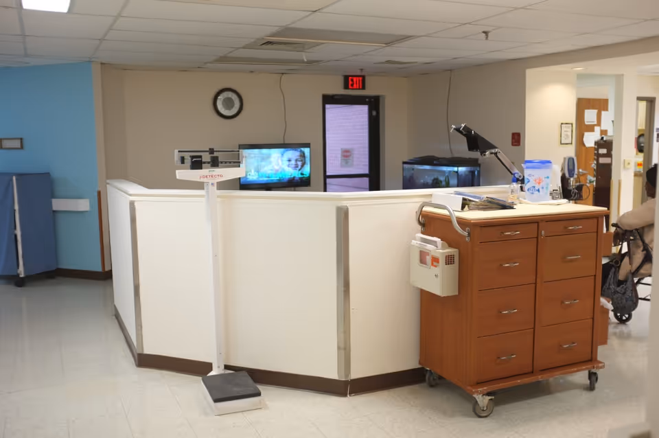 Interior view of a nursing station area in an assisted living facility. A white scale stands in front of a white counter with a clock on the wall behind it. A wooden medical cart with drawers and a lamp is positioned to the right. A television screen is mounted on the wall behind the counter, and a door with an exit sign is visible in the background. A person in a wheelchair is partially visible on the right side.