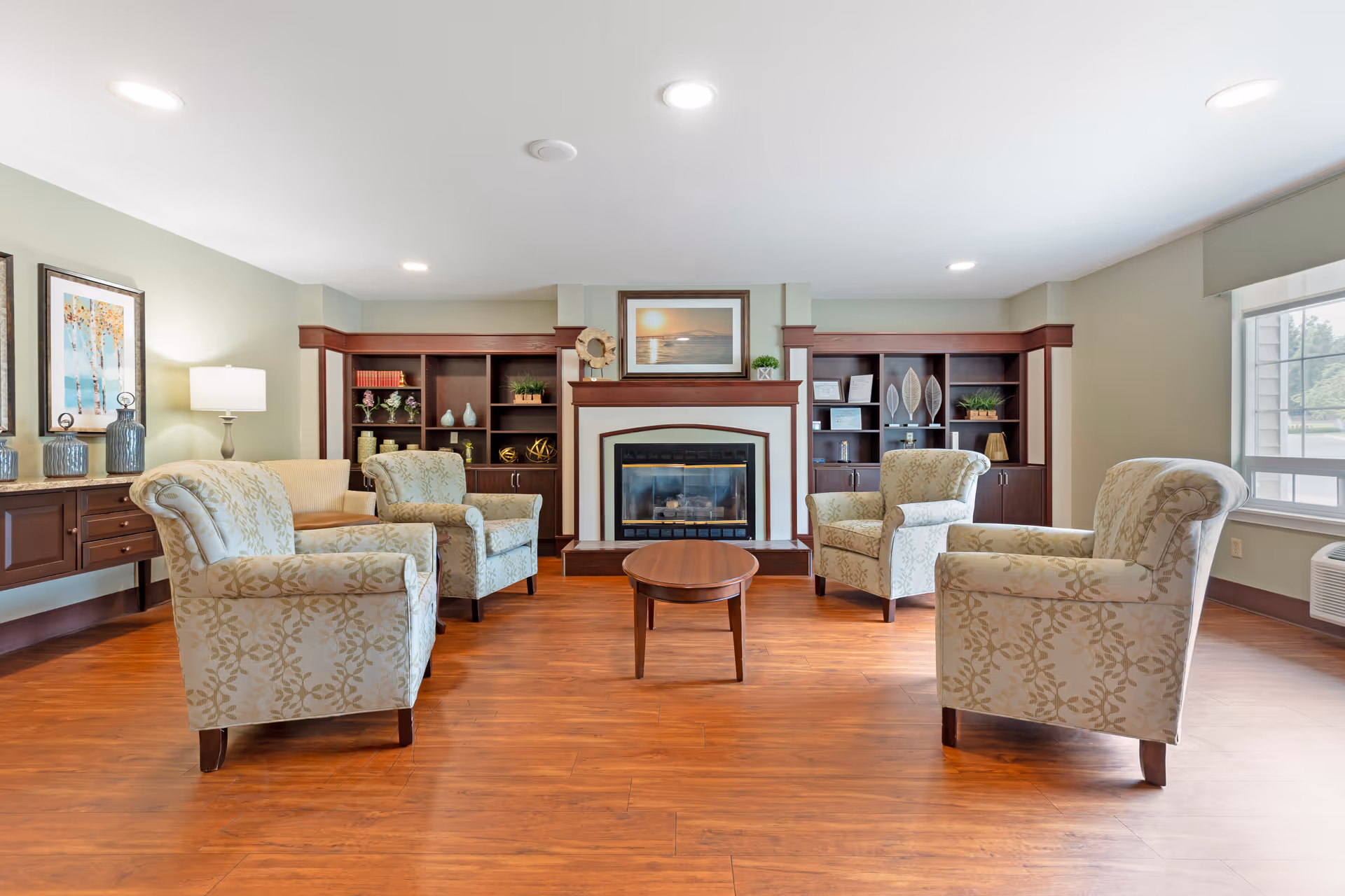 A cozy living room area with four patterned armchairs arranged around a small wooden coffee table. The room features a fireplace with a framed picture above it, flanked by built-in wooden shelves decorated with plants, books, and ornaments. There is a large window on the right side letting in natural light, and a wooden sideboard with a lamp and decorative vases on the left. The floor is wooden and the walls are painted light green.