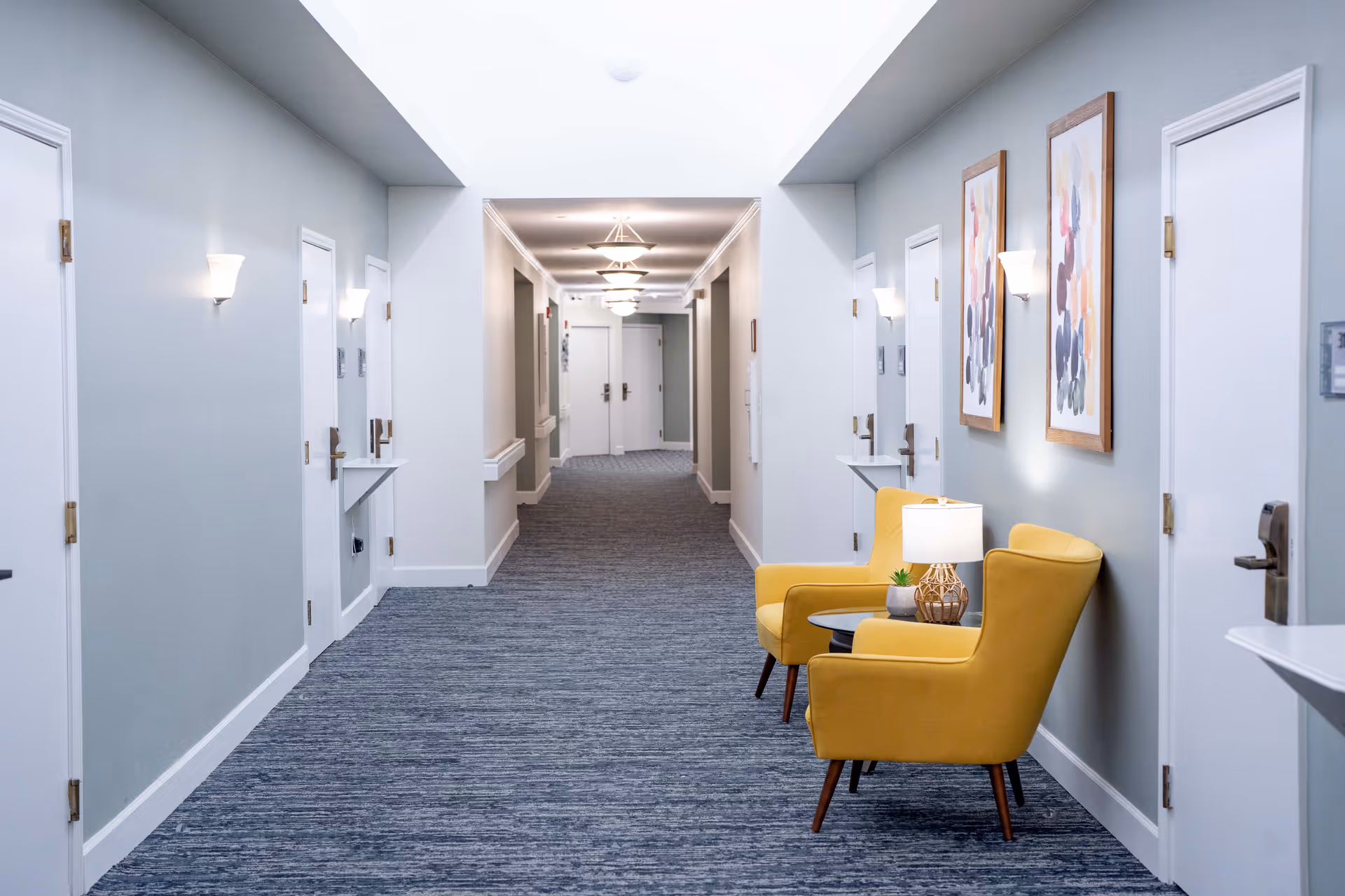 A well-lit hallway in a senior living facility with multiple white doors on both sides, blue carpet flooring, and light gray walls. On the right side, there are two yellow armchairs with a small round table between them holding a lamp and a small plant. Two abstract paintings hang on the wall above the chairs.