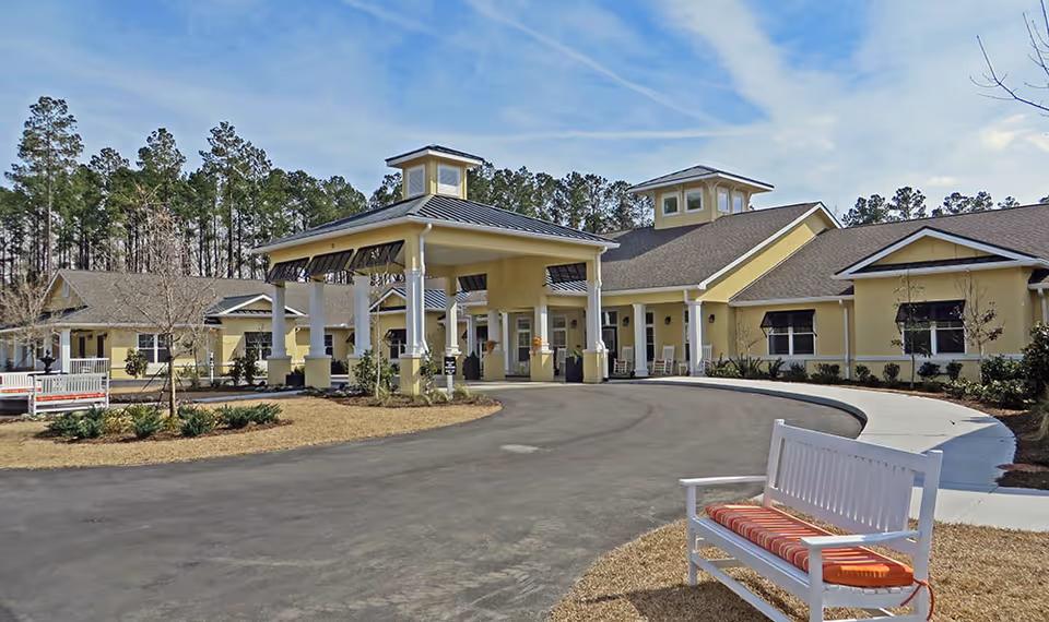 Exterior view of The Addison of Bluffton senior living facility showing a large yellow building with a covered entrance, surrounded by trees and a paved driveway. There is a white bench with an orange cushion in the foreground on a grassy area.