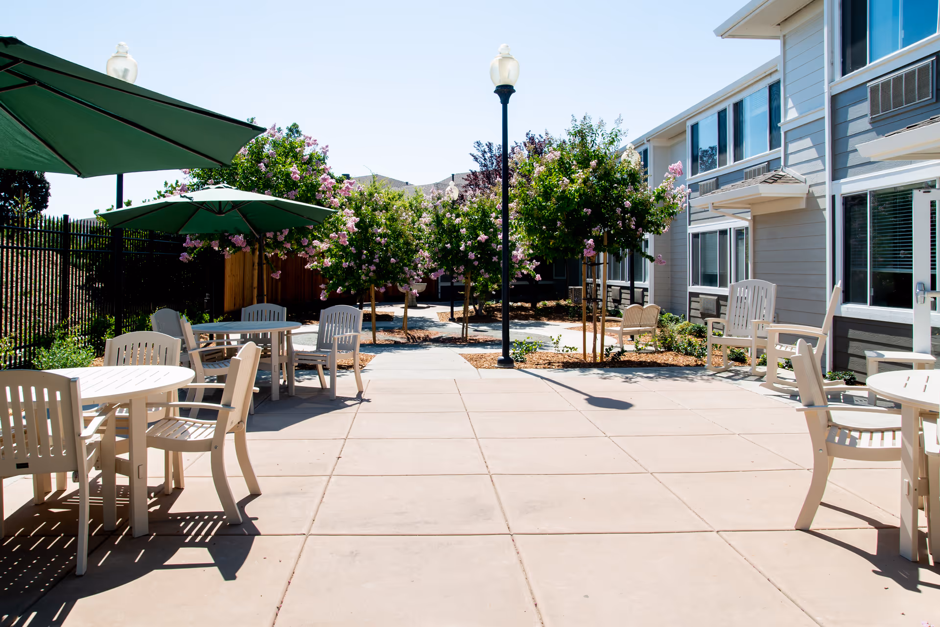 Outdoor patio courtyard with round tables, chairs, green umbrellas, flowering trees, and the exterior of a senior living building.