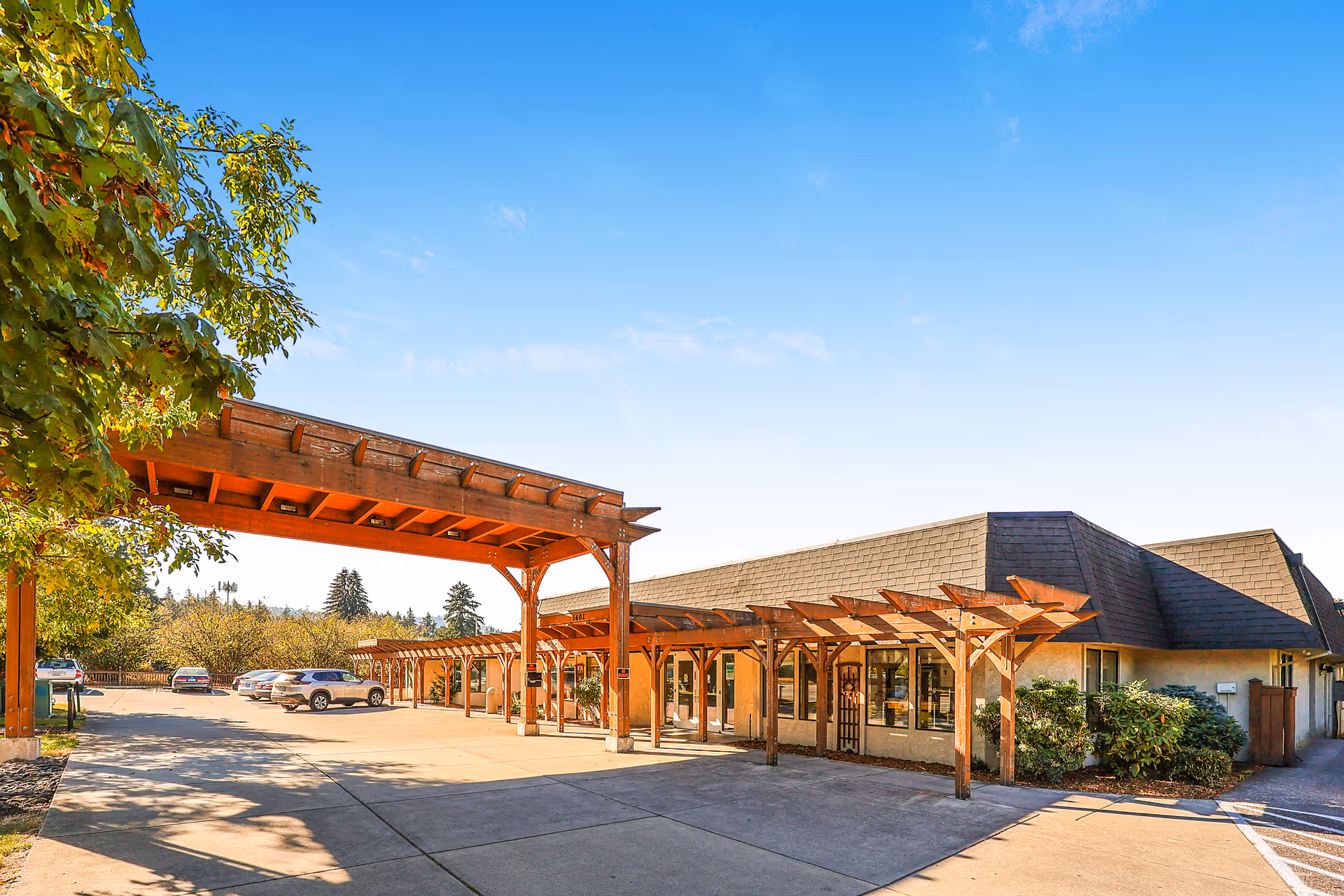 Exterior view of Cascade Terrace Post Acute facility showing a single-story building with a dark shingled roof and wooden pergolas along the entrance. Several cars are parked in the parking lot under a clear blue sky with some trees visible on the left side.