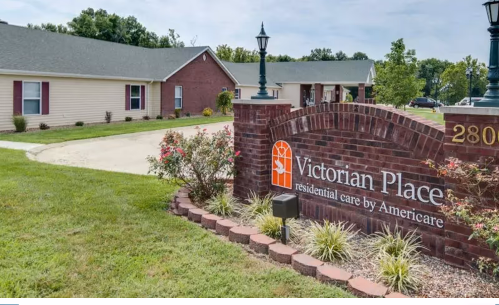 Exterior view of Victorian Place of Washington Senior Living facility showing a brick sign with the facility name and logo, surrounded by landscaped grass, plants, and flowers. The building with beige siding and red brick accents is visible in the background under a partly cloudy sky.