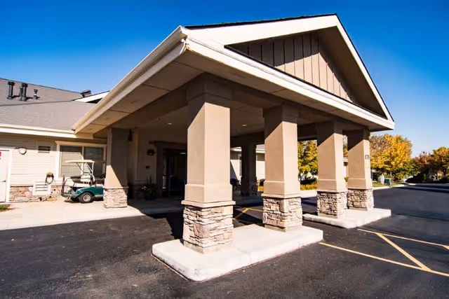 Front entrance of a senior living facility with a covered drop-off area supported by four large columns with stone bases, a golf cart parked to the side, and a clear blue sky overhead.