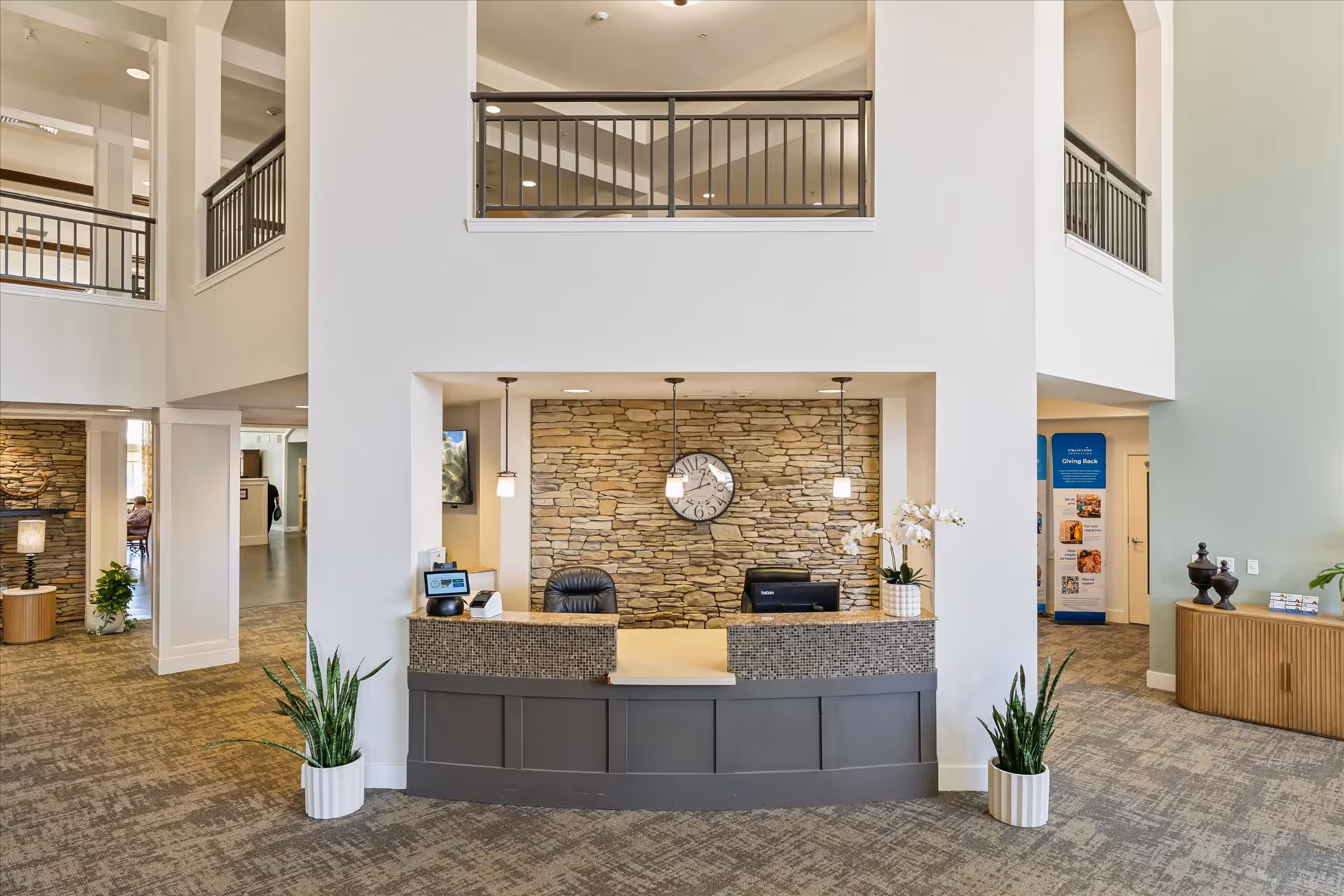 Reception area with a front desk featuring a stone accent wall and a clock above it. The desk has two pendant lights hanging from the ceiling, a computer, a chair, and decorative plants on either side. The space has a high ceiling with a balcony railing visible on the upper level and carpeted flooring.