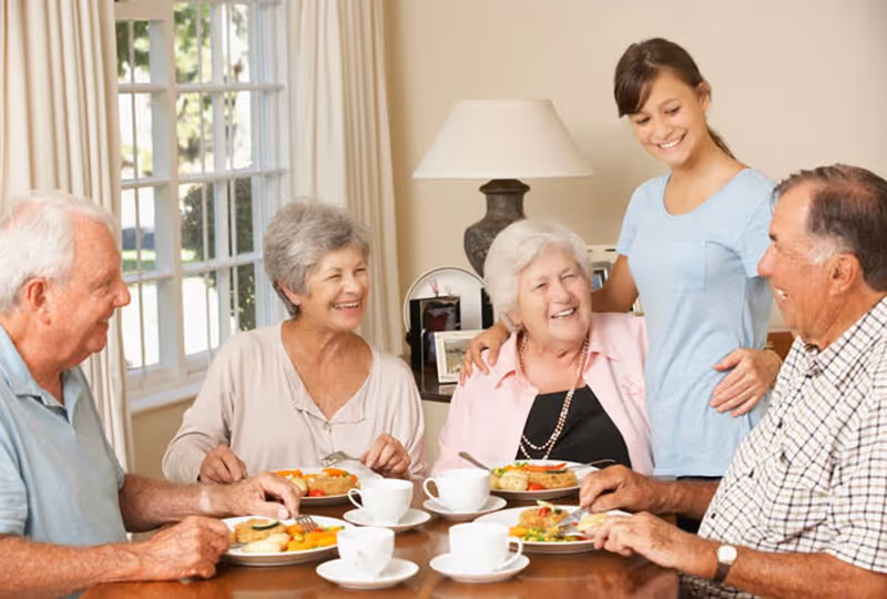 Four elderly people sitting around a dining table enjoying a meal together, with a younger woman standing behind them smiling and placing her hands on two of the elderly individuals' shoulders. The room is well-lit with a window and a lamp in the background.