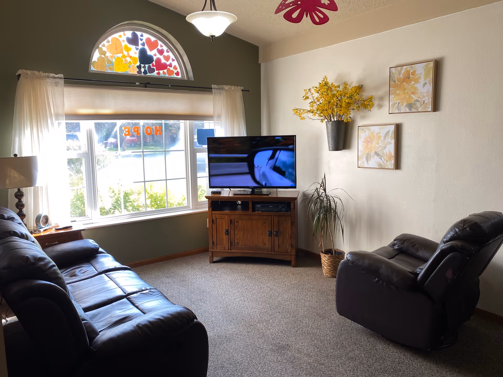 A cozy living room with a large window decorated with colorful heart-shaped stickers and the word HOPE. The room features a dark leather sofa and recliner, a wooden TV stand with a flat-screen TV, a floor lamp, and wall decorations including yellow floral paintings and a vase with yellow flowers.