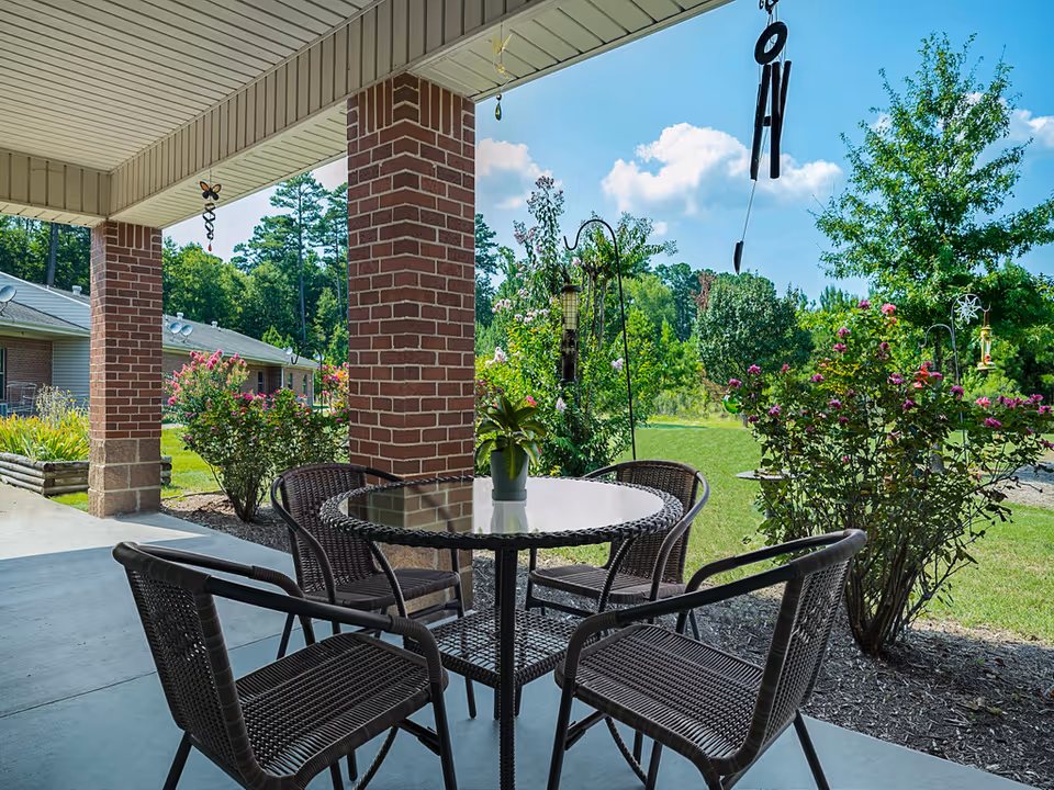 Outdoor covered patio area with a round glass-top table and four wicker chairs. The patio has brick columns and overlooks a garden with flowering bushes and green trees under a blue sky with some clouds. Wind chimes hang from the patio ceiling.