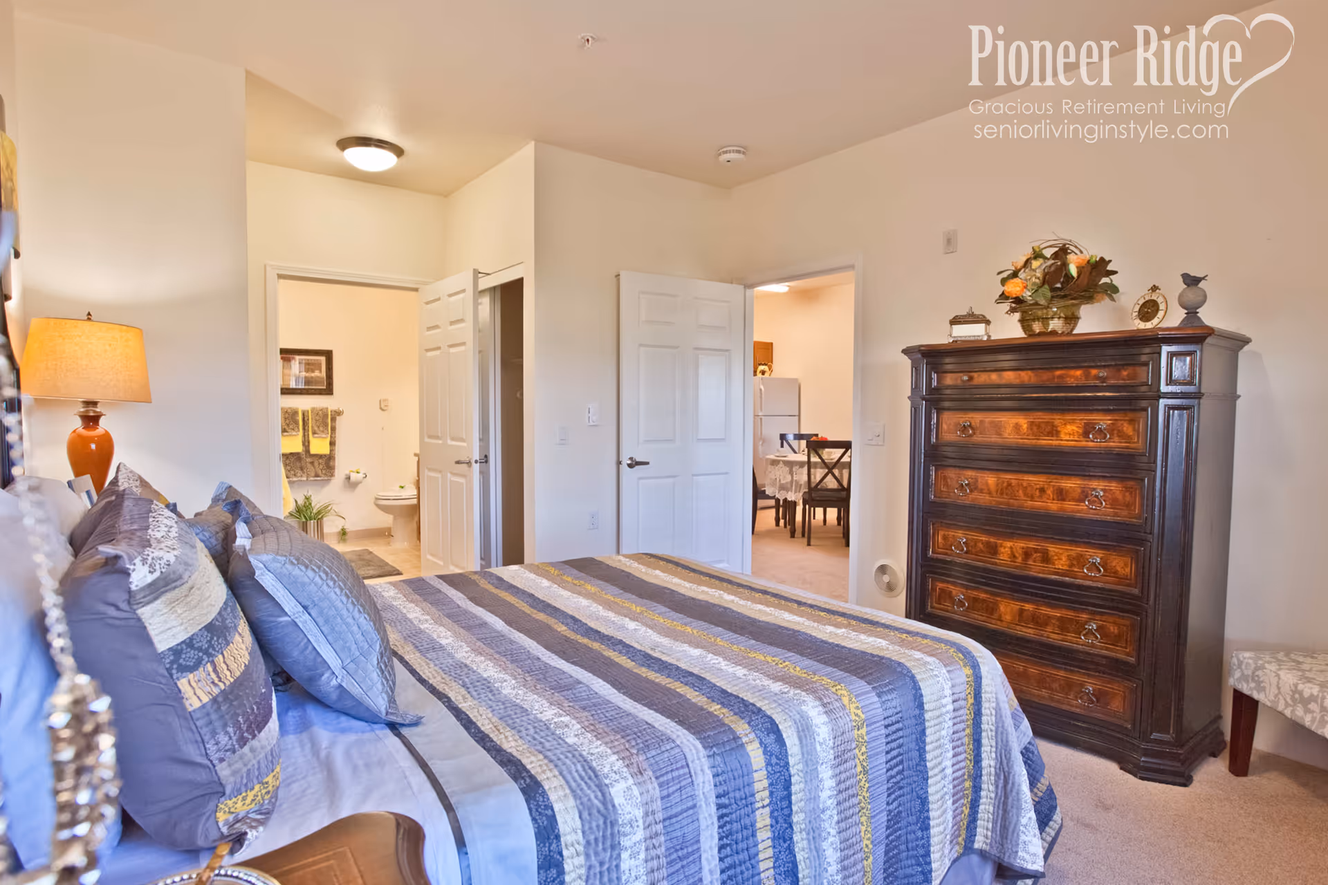 A cozy bedroom with a bed covered in a striped quilt in shades of blue, gray, and yellow. There are multiple pillows on the bed and a bedside lamp with an orange base. The room has beige walls and carpet. A dark wooden chest of drawers with decorative items on top is against one wall. Two open doors reveal a bathroom with towels and a toilet, and a dining area with a table and chairs. The image has a watermark in the top right corner that reads 'Pioneer Ridge Gracious Retirement Living seniorlivinginstyle.com'.