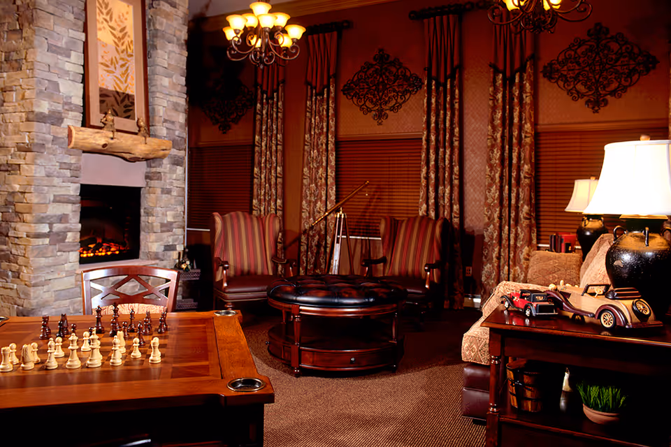 Cozy common living room with a stone fireplace, chessboard on a table, upholstered chairs, ottoman, and lamps.