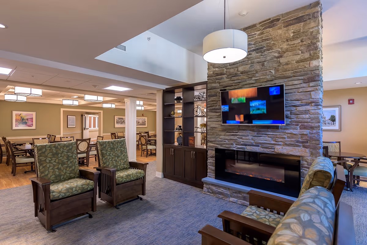 A cozy senior living facility common area with two green patterned armchairs facing a stone fireplace with a mounted flat-screen TV above it. Behind the chairs is a dark wood shelving unit, and in the background is a dining area with multiple tables and chairs, soft lighting, and framed artwork on the walls.