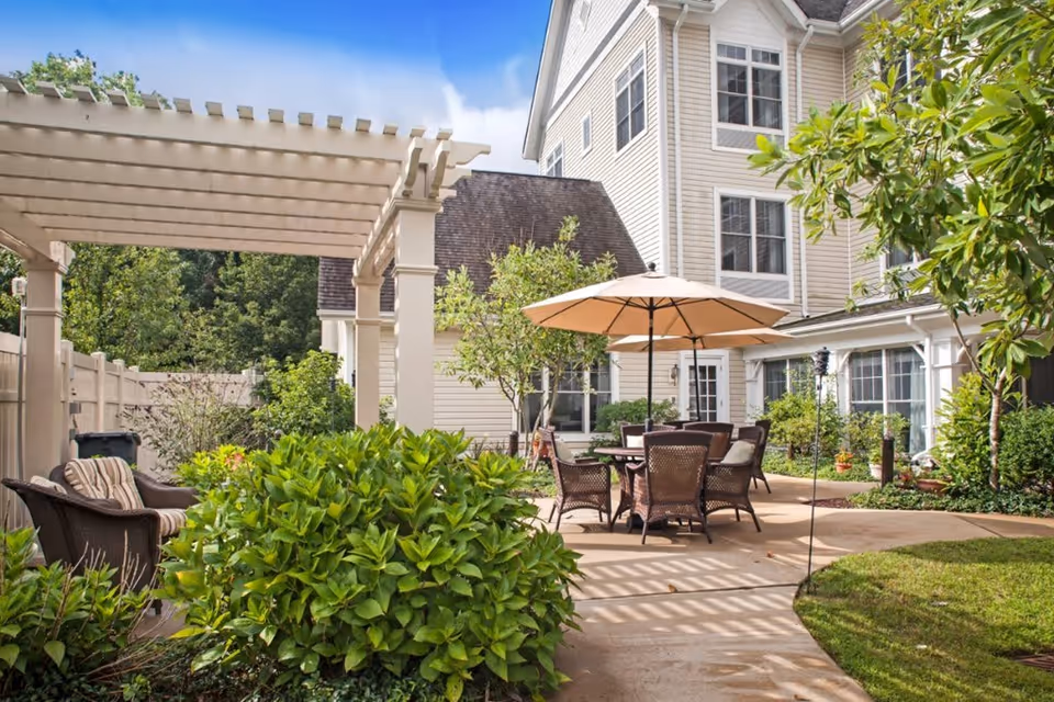 Outdoor patio area at a senior living facility with a beige pergola, wicker chairs with cushions, a round table with an umbrella, surrounded by green bushes and trees, and a multi-story beige building in the background under a blue sky with some clouds.