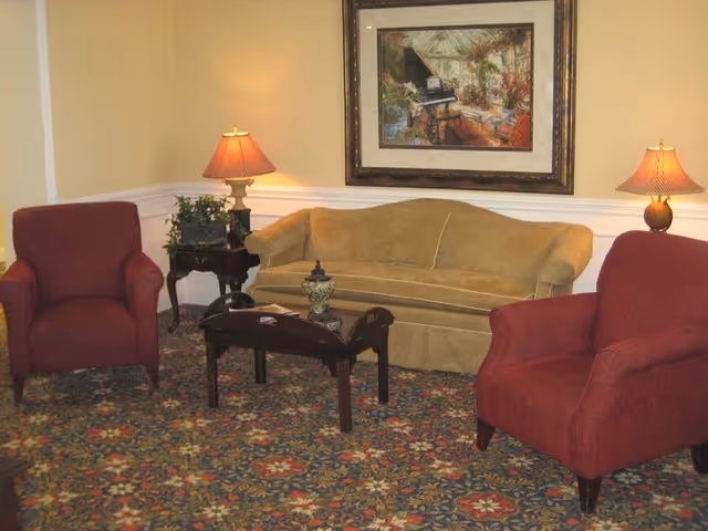 A cozy living room area with a mustard yellow sofa, two red armchairs, a dark wooden coffee table, two table lamps with red shades on side tables, a potted plant, and a framed painting of a piano and sunlit room on the wall.